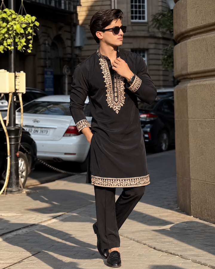 A man wearing a black long kurta with beige and gold embroidery on the neckline and cuffs, paired with black trousers, walking on a city sidewalk.